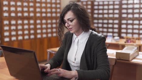 Lächelnde-Studentin-Tippt-Auf-Ihrem-Laptop-An-Einem-Holzschreibtisch-In-Der-Bibliothek