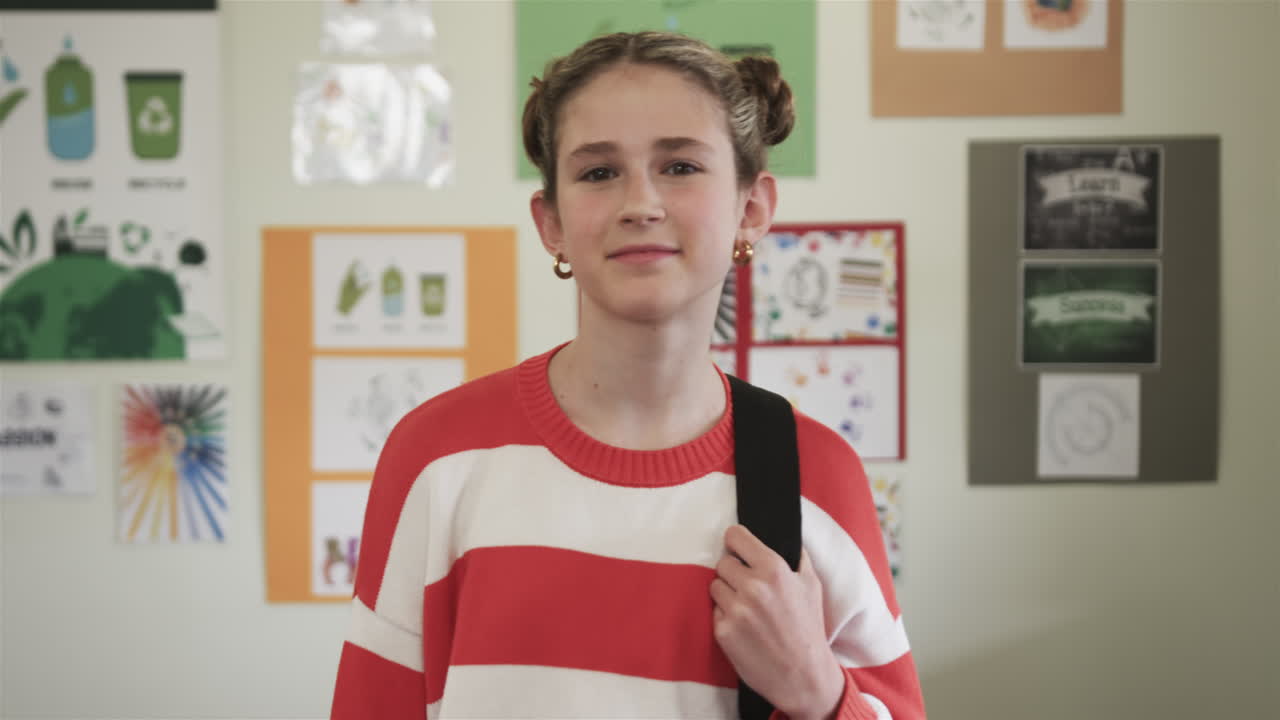 In classroom, girl with backpack smiling, standing near colorful posters, at school