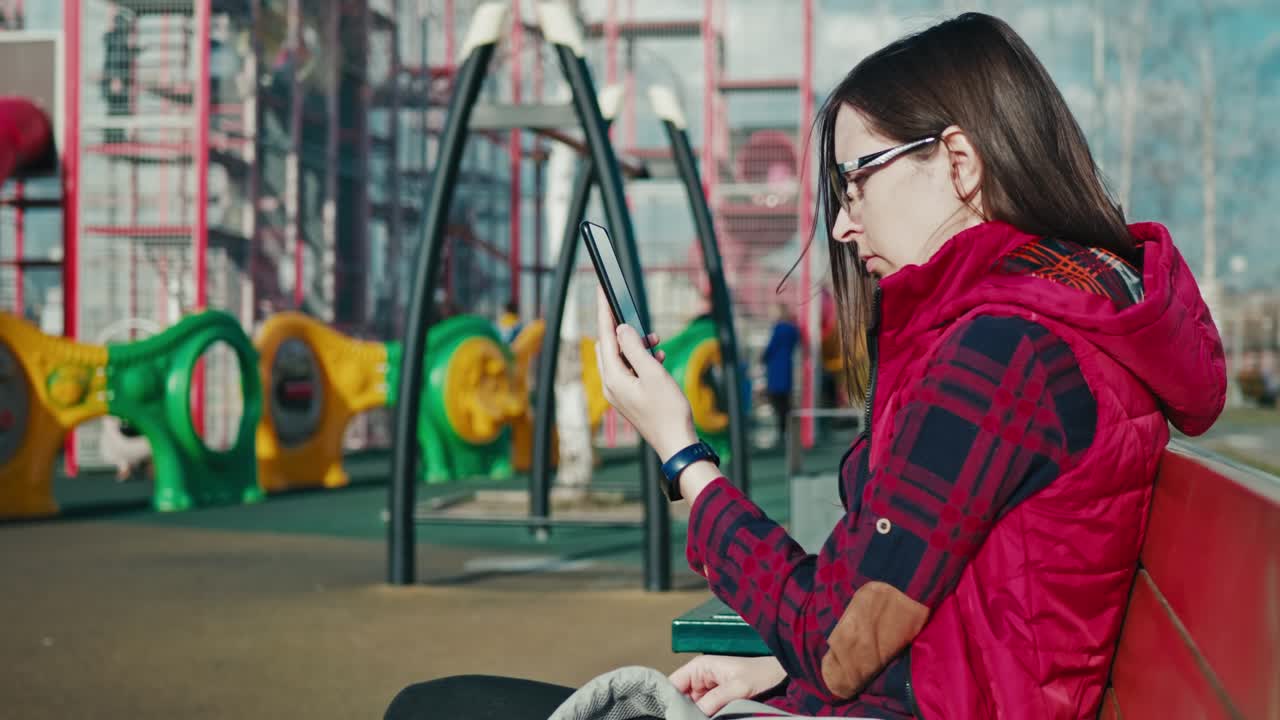 mother uses smartphone and watching children playing on playground.