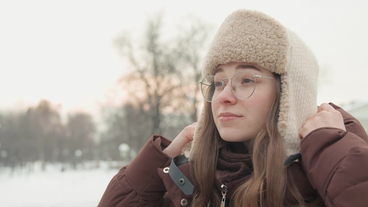 Three quarter chest up portrait of young woman smiling while adjusting fur lined trapper hat in snowy park at dusk, with soft light, cold air, blurred trees and relaxed seasonal mood