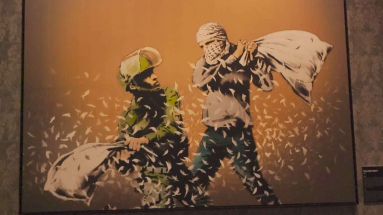 An oil painting or stencil art made by Banksy showing two soldiers fighting with pillows. An Israeli and Palestinian soldier fighting with pillows that are broken and feathers fly out. Framed art