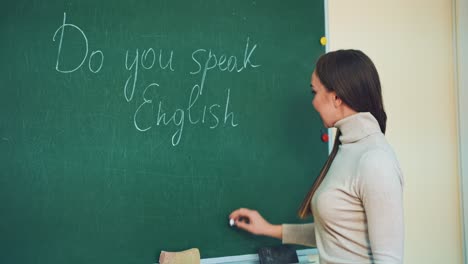attractive teacher of english writes on a green board with a chalk. beautiful long-haired woman is writing question tag on the blackboard and smiles to camera in class.