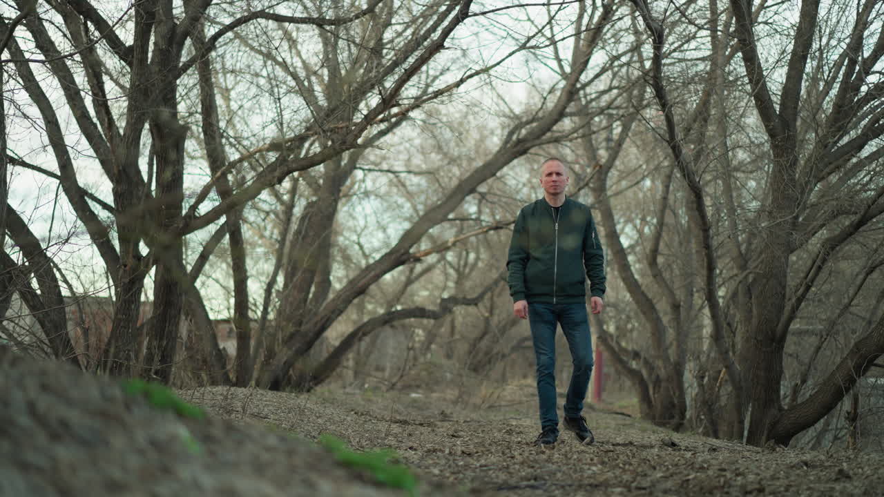 un hombre camina a través de un denso bosque con árboles desnudos a principios de la primavera, con una chaqueta negra, pantalones vaqueros y lona
