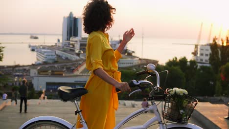 attractive brunette woman in a long yellow dress during the dawn standing by her city bicycle holding its handlebar with flowers