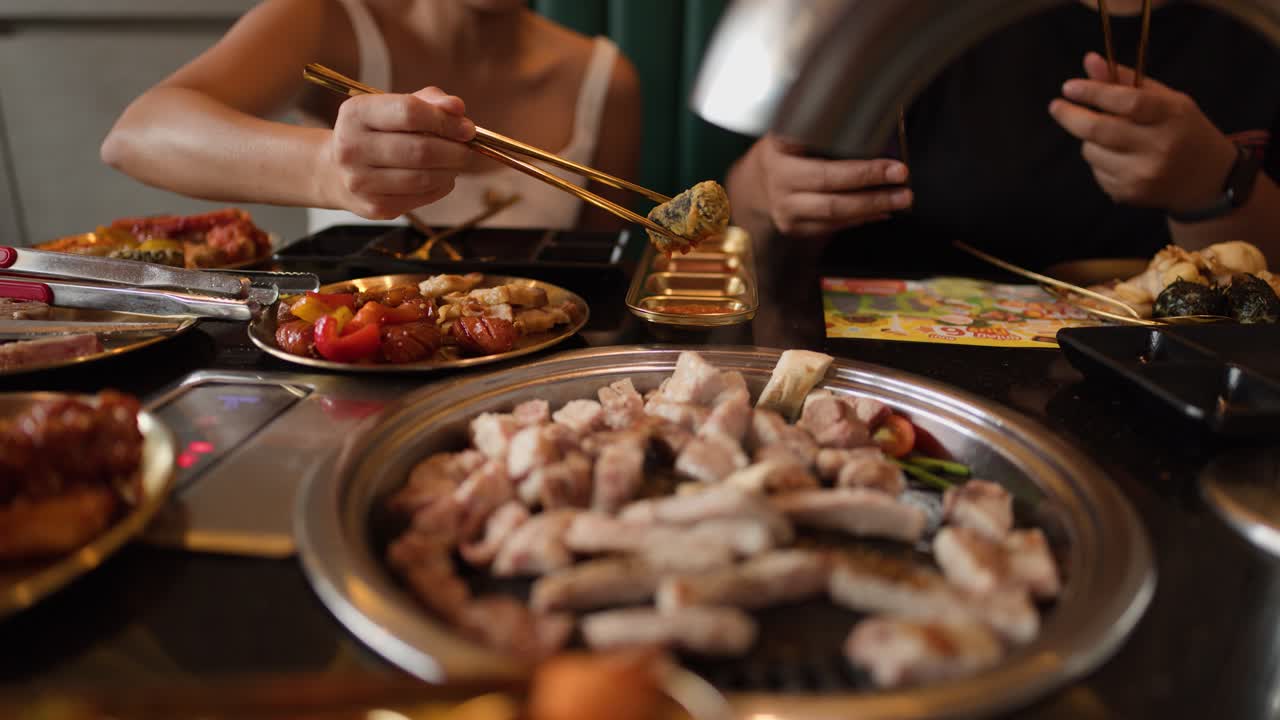 Two people grill meat and dip food in sauce, enjoying Asian barbecue restaurant ambiance