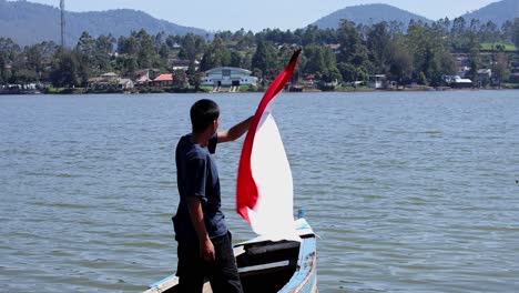 young man standing on traditional wooden boat and waving indonsia flag in lake