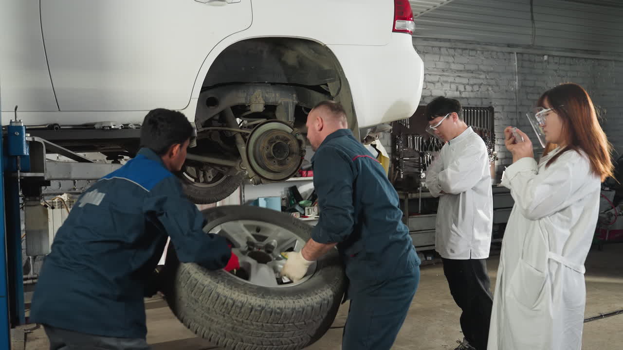 estudiante en bata de laboratorio tomando una foto de un mecánico trabajando en un coche elevado en un taller de reparación de automóviles mientras otro estudiante observa con los brazos cruzados