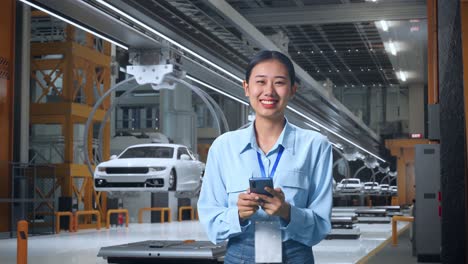 trabajadora profesional asiática de pie con su smartphone en una fábrica de automóviles ev, instalación industrial de alta tecnología, está mirando a la cámara con una sonrisa