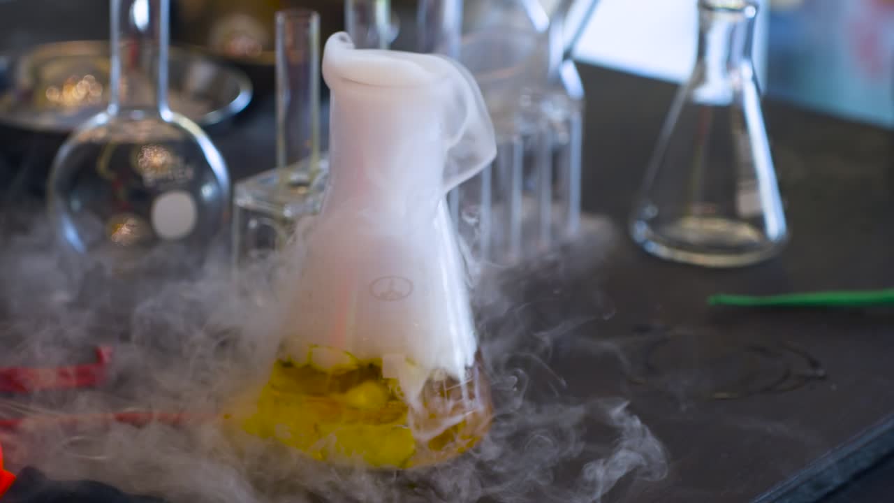 Primo piano di un esperimento scientifico che mostra una fitta nebbia bianca di ghiaccio secco che fuoriesce violentemente da un liquido giallo in una beuta di Erlenmeyer, circondata da vetreria da laboratorio e strumenti su un banco di lavoro.