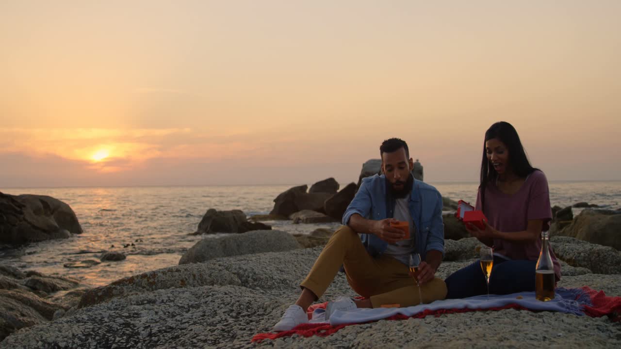 feliz pareja de raza mixta dando regalos el uno al otro durante la puesta de sol en la playa 4k