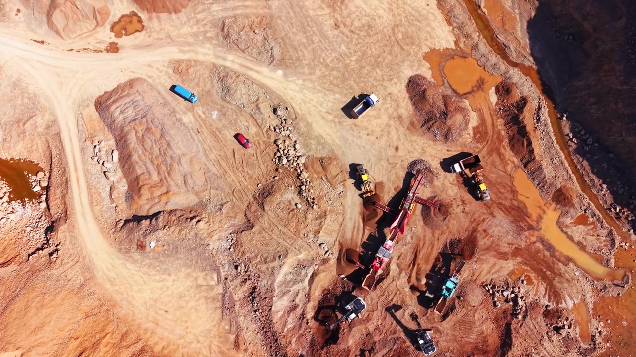 Quarry trucks and crusher machinery, Bulgaria. Overhead view of colorful vehicles on red dirt roads beside orange mineral ponds.