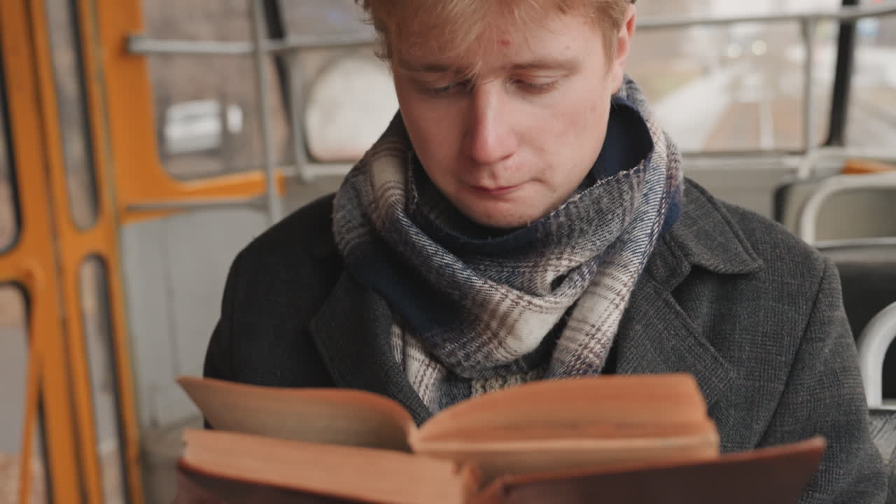 Junger weißer Kreativer liest in der Straßenbahn, kuscheliger Schal, Wollmantel, vertieft in ein altes Hardcover-Buch, langsames Umblättern der Seiten, Nahaufnahme der Hände und nachdenklicher Blick, urbane Pendleratmosphäre, kontemplative Leseszene für einen Schriftsteller.