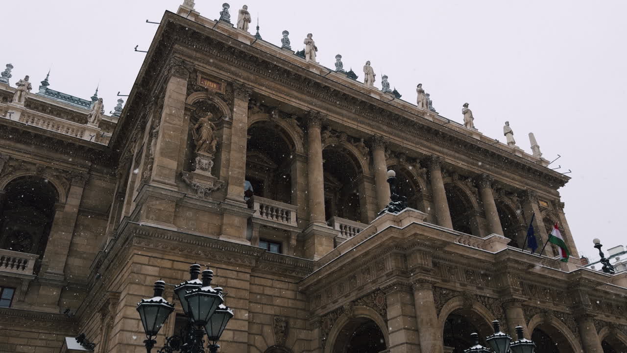 Vista ampla da Ópera Estatal Húngara em Budapeste durante uma nevasca. Arquitetura europeia histórica, colunas de pedra, estátuas e atmosfera urbana de inverno.