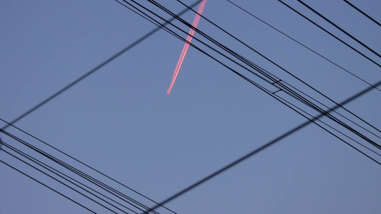 Silhouette powerlines with pink aircraft vapor trail against blue sky, abstract
