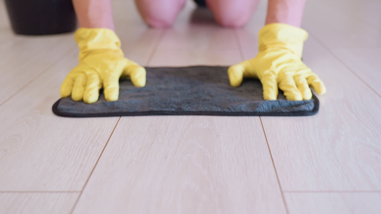 Close up of home serviced cleaner kneeling with yellow gloves gently moving rag across wooden floor, focusing on hygiene, sanitation, household maintenance, and careful attention to domestic cleaning