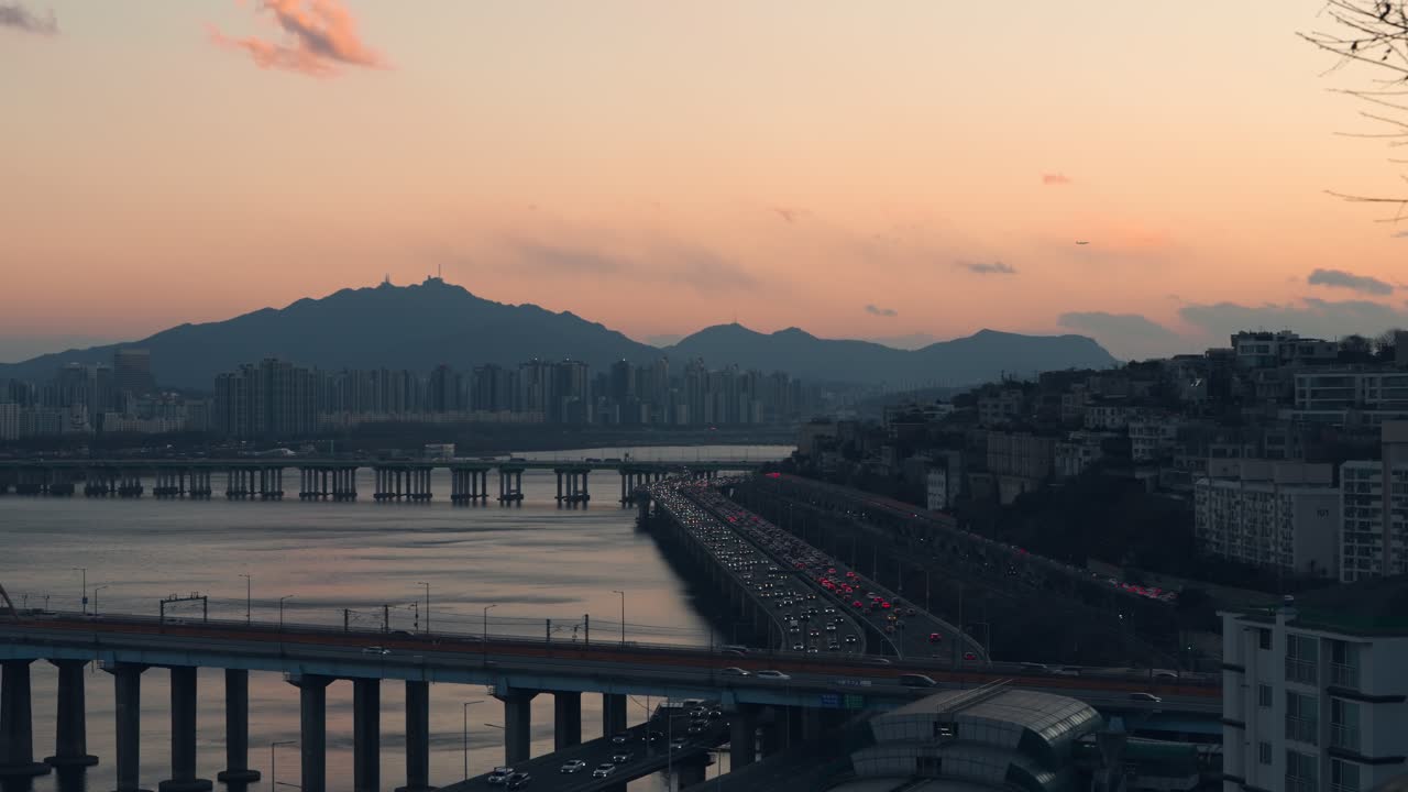 Ripresa aerea del traffico intenso sull'autostrada Gangbyeonbuk-ro e sul ponte Dongho sul fiume Han al crepuscolo, con skyline della città e silhouette di montagne a Seoul, Corea del Sud.