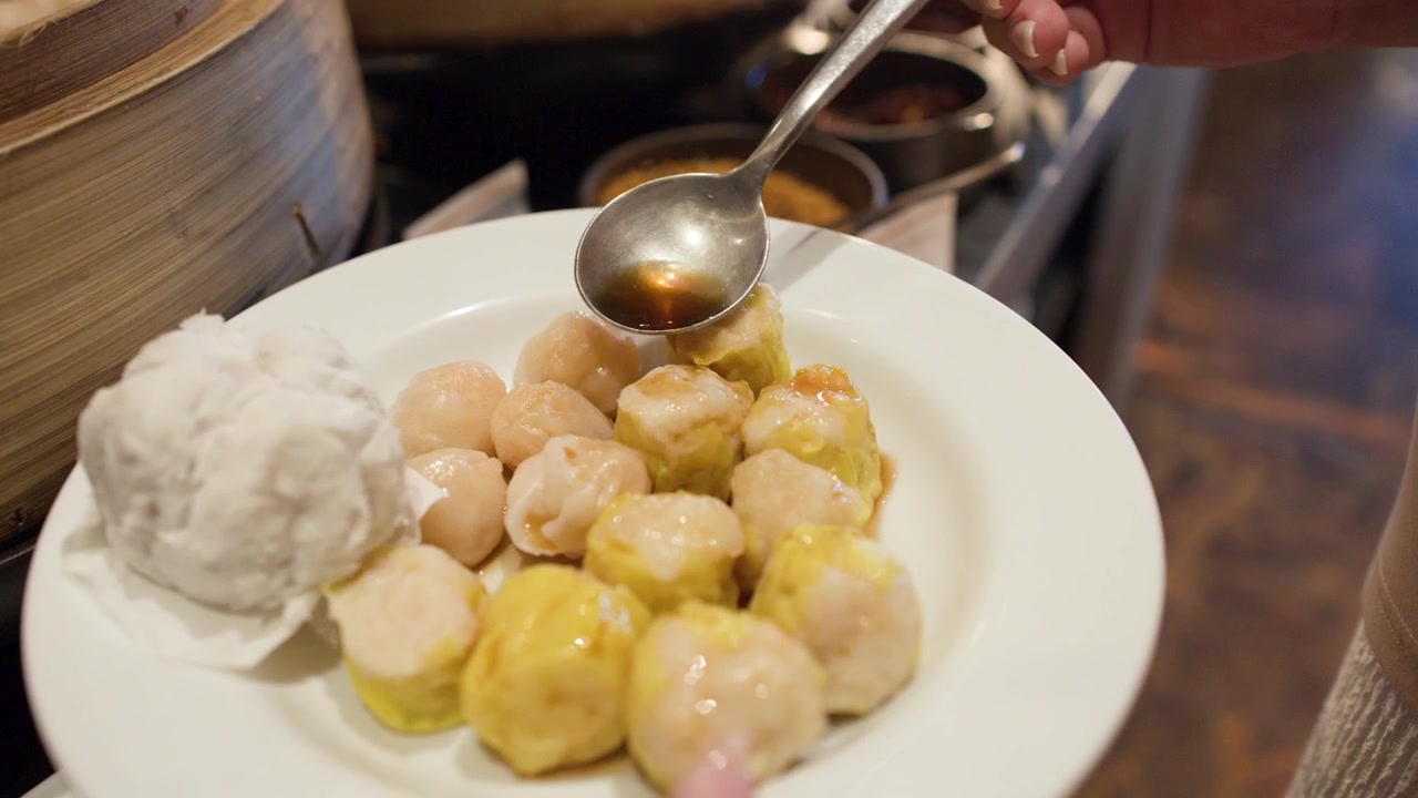 A person pours soy sauce from a spoon onto assorted dim sum on a plate