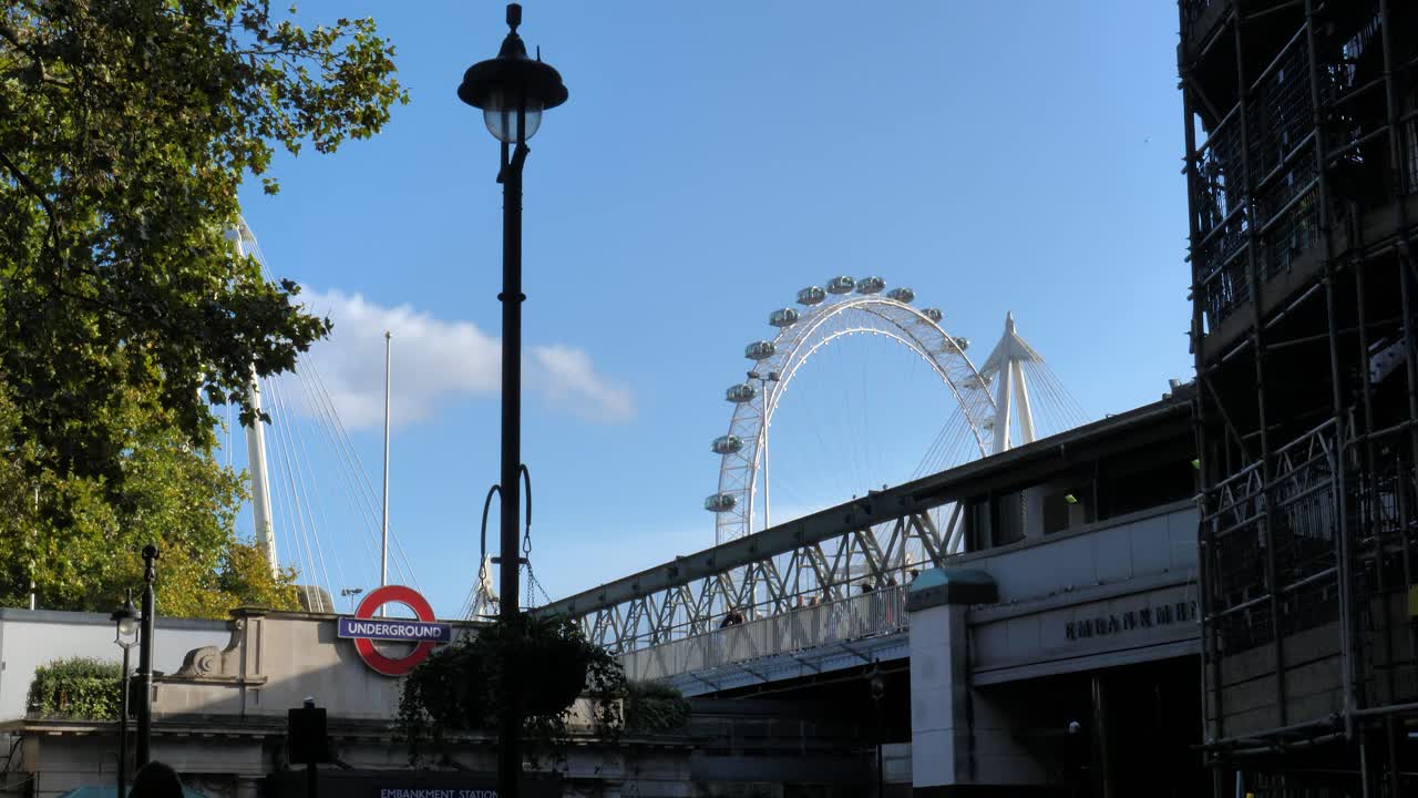 Iconic elements in London: the underground station, a street light in the foreground and the London Eyes, the Big Wheel on the river Thames, in the background. Some turists are walking on the bridge.