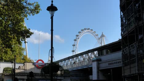 iconic elements in london: the underground station, a street light in the foreground and the london eyes, the big wheel on the river thames, in the background. some turists are walking on the bridge.