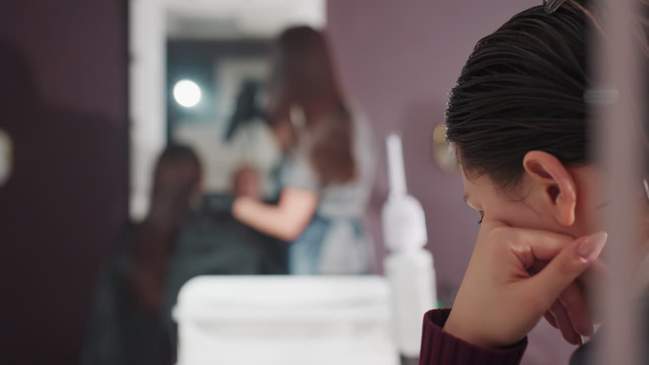 젖은 머리를 하고 턱에 손을 괸 채 골똘히 생각하는 고객, 샴푸 및 헹굼 단계에서 고민하는 백인 고객, 배경에서 작업 중인 스타일리스트, 섬세한 클로즈업, 차분한 분위기와 은은한 팔레트