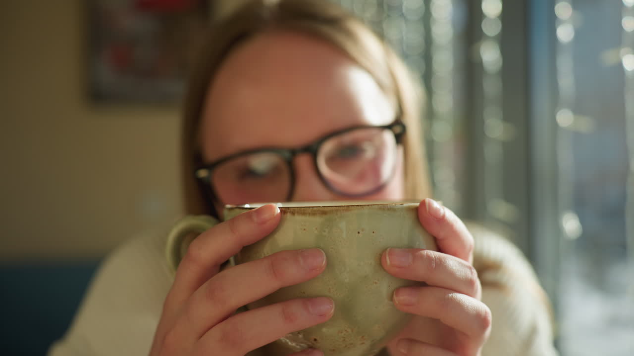 Close up of stylish woman wearing glasses, gently holding ceramic mug with both hands while taking tea by large window decorated with string lights