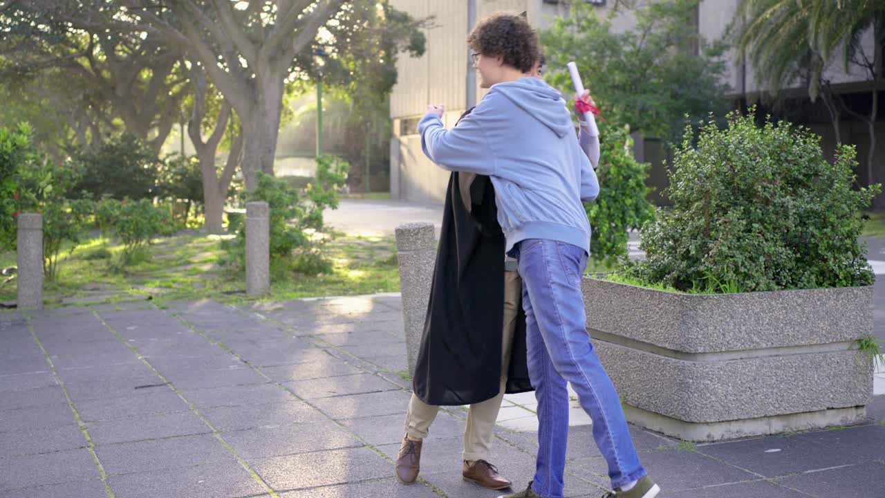 Verschiedene männliche Freunde auf dem Campus absolvieren in Robe, halten Diplome, während ein Freund sich nähert, umarmt und spricht.