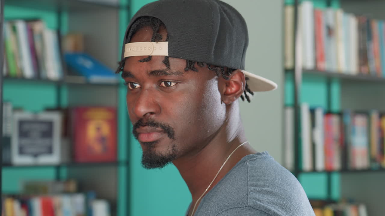 Student in casual clothing and backward cap standing in library, lifting head slightly with thoughtful gaze, surrounded by shelves of books in colorful background