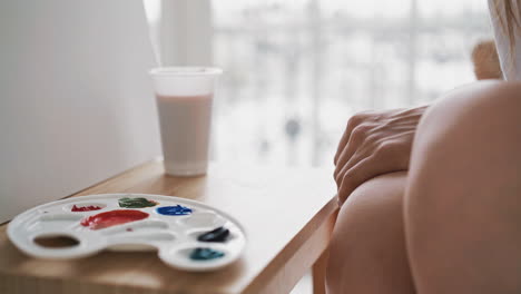 Artist-sits-near-palette-with-paints-and-cup-on-wooden-easel