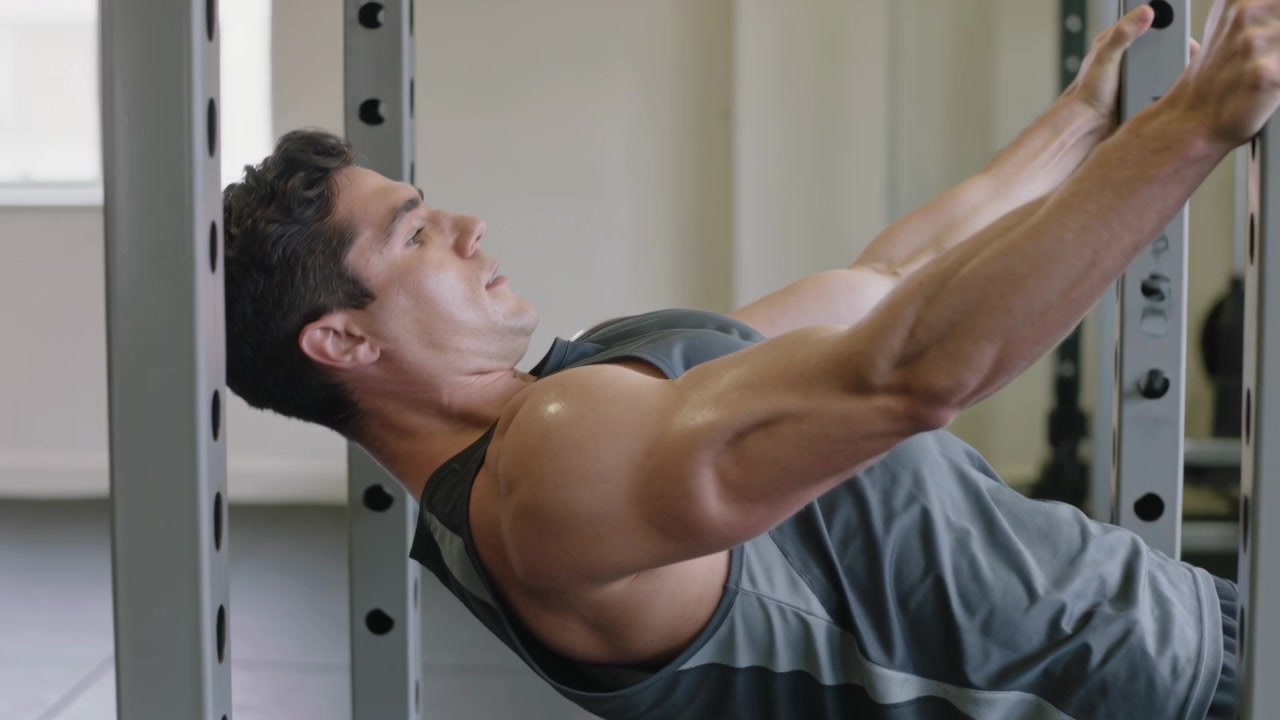Pulling fit male wearing grey tank starting from arms extended under rack-bar at gym, toning back