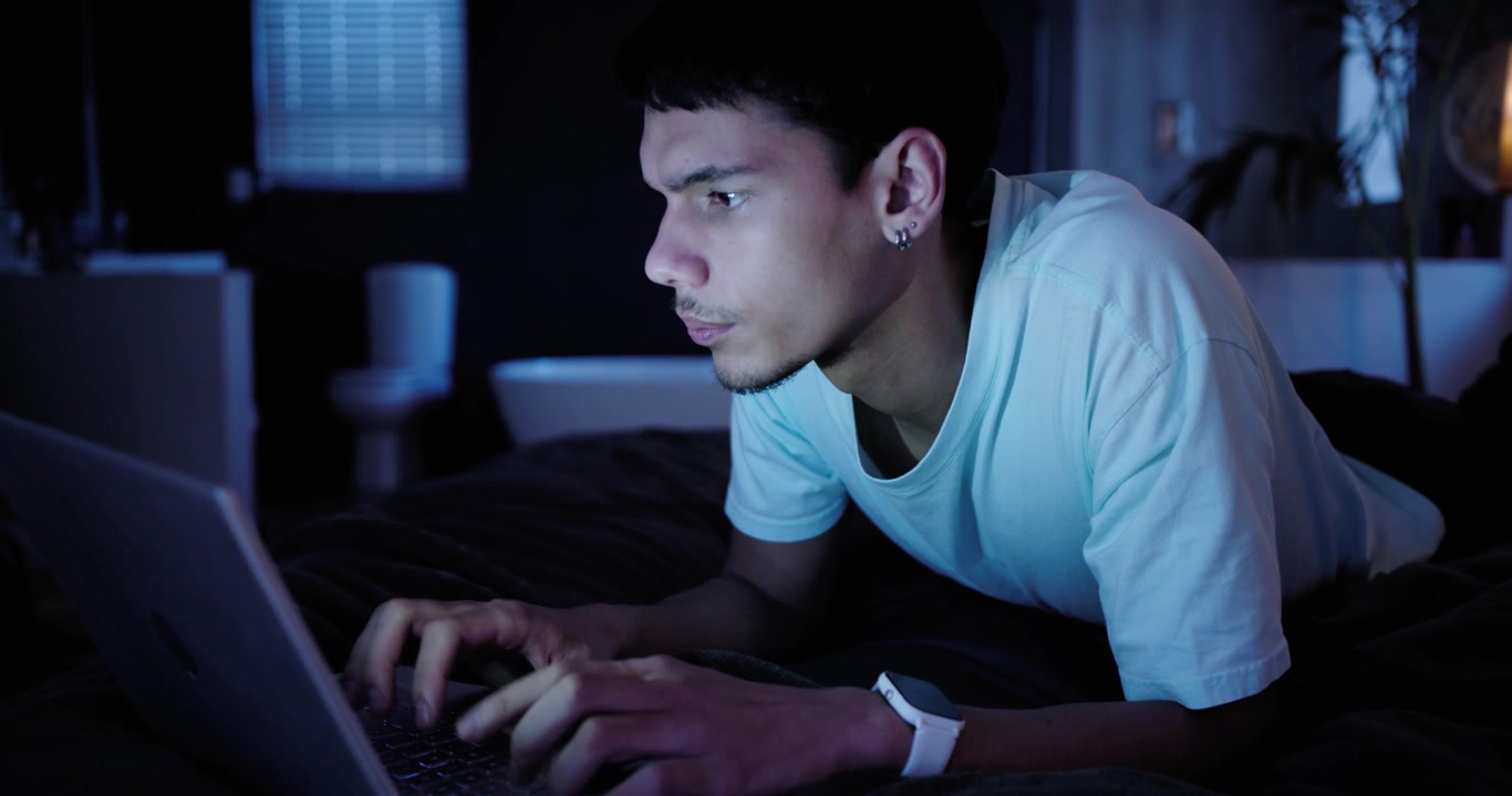 Teenage male lying on stomach on bed, laptop blue glow drawing focus, studying and typing