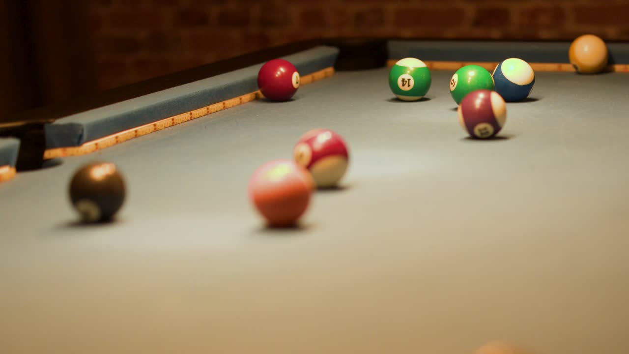 Hand strikes cue ball, scattering pool balls across indoor billiard table under warm, low lighting