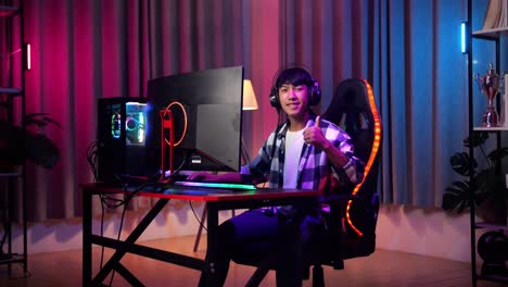 adolescente asiático sonriendo a la cámara y mostrando el pulgar hacia arriba mientras juega videojuegos en la computadora