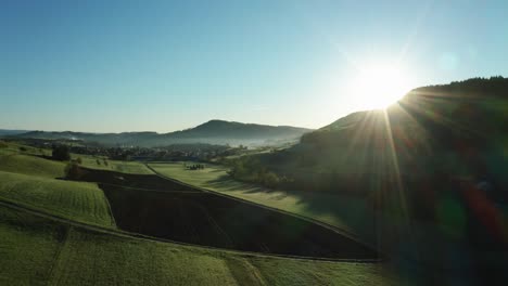 aerial-drone-view-of-morning-hour-over-peaceful-countryside-with-green-and-agricultural-fields,-sun-shining-directly-into-camera,-switzerland