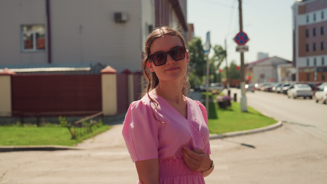Sonnengefluteter Straßenspaziergang mit kaukasischer Frau in Pink. Modebloggerin geht in fließendem Chiffonkleid und Sonnenbrille in der Innenstadt spazieren, Zebrastreifenszene, warmes Nachmittagslicht, offenes Lächeln, Ladenfronten von Boutiquen, sanft