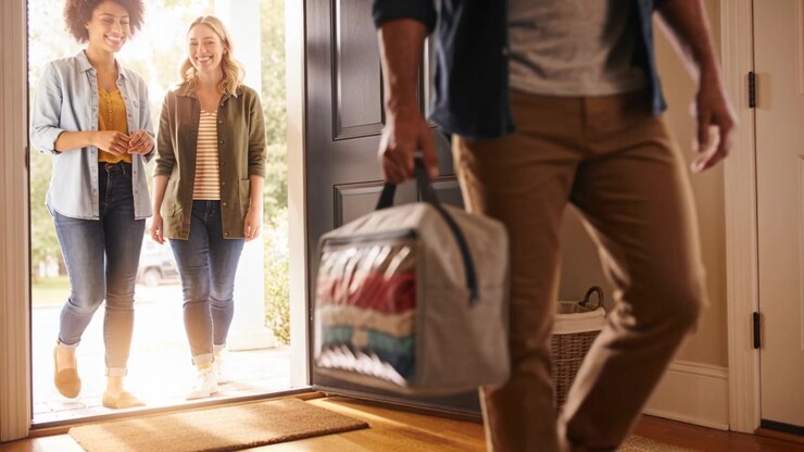 Door opening, man in dark shirt holding bag of textiles stepping into foyer, women smiling follow
