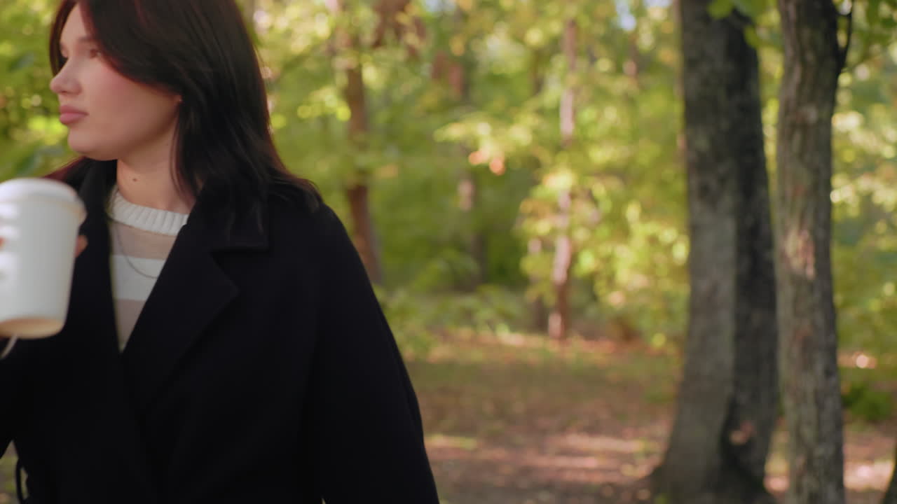 숲길을 거닐면서 머그잔에 담긴 따뜻한 음료를 마시고, 가을 낙엽을 들고, 긴 코트를 입고, 부드러운 햇빛 아래에서 여유로운 걸음을 걷는, 평화로운 자연 속에서 아늑한 분위기를 내는 천진난만한 소녀