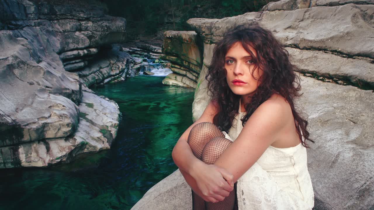 4K Cinemagraph - seamless video loop of a beautiful young brunette model woman in a white dress sitting at a gorge by a blue mountain river. The water is rushing through naturally formed rocks