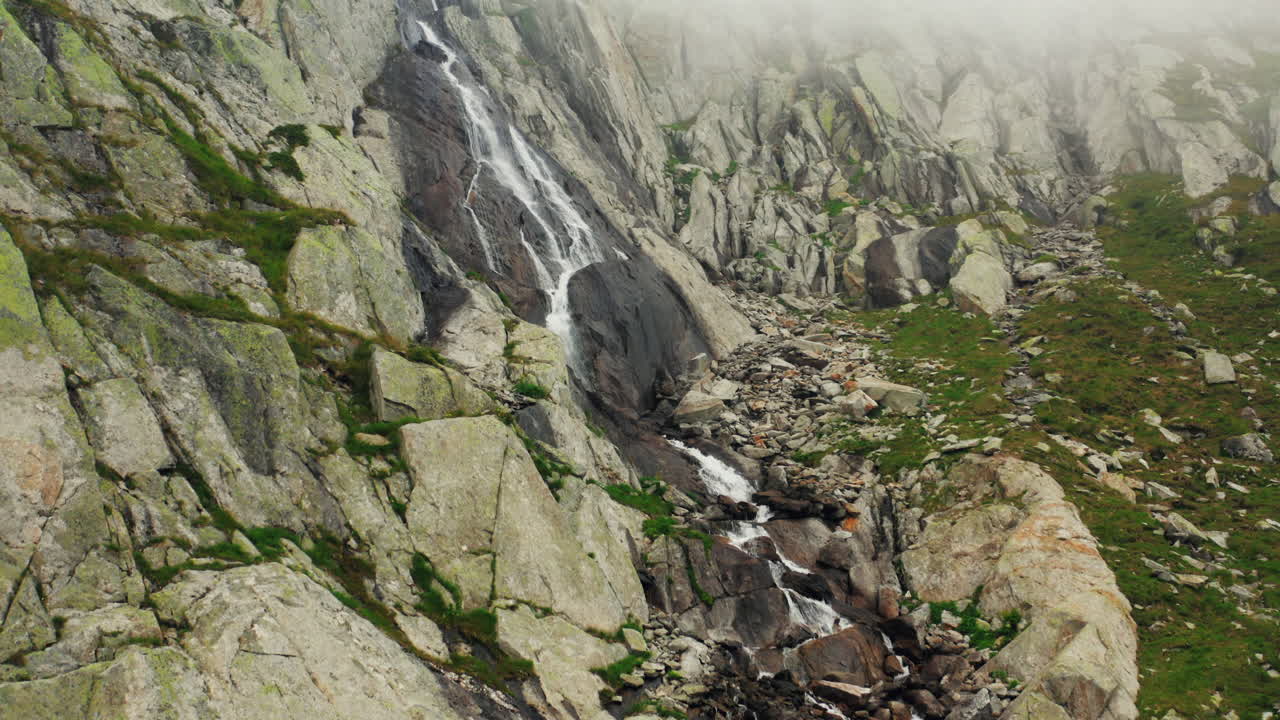 Mountain waterfall cascading down gray rocky cliff in misty alpine landscape. sparse vegetation. Drone push in, tilt up.