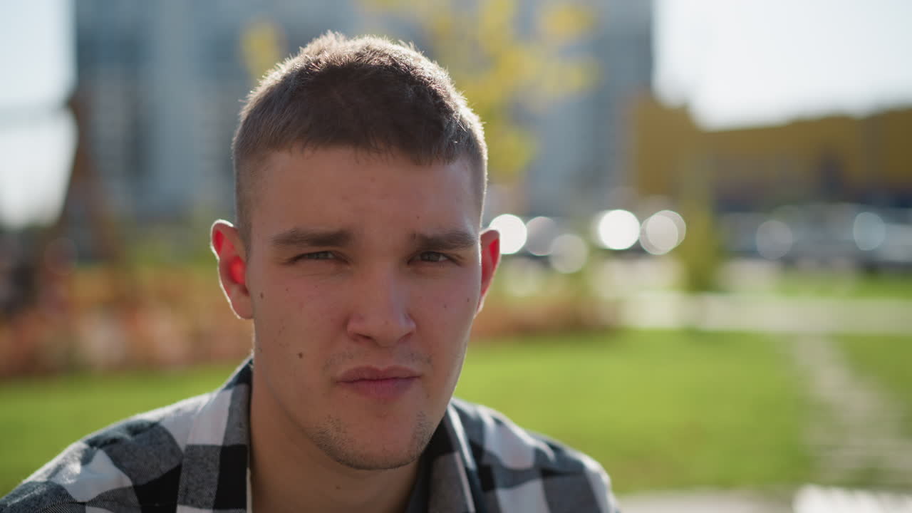 portrait view of fair skinned man with short hair wearing checkered shirt staring into camera under bright sun with blurred background of greenery, pathway, and parked cars in urban outdoor setting