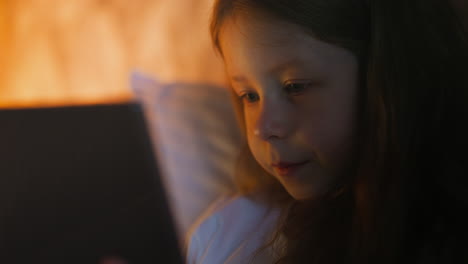 una niña pequeña habla mirando la tableta en la cama en una habitación sombreada