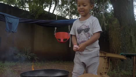 a young mixed raced boy throws a sausage on a frying pan over a camp fire in his backyard