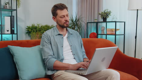 Young-man-freelancer-sitting-on-sofa-closing-laptop-pc-after-finishing-work-in-living-room-at-home