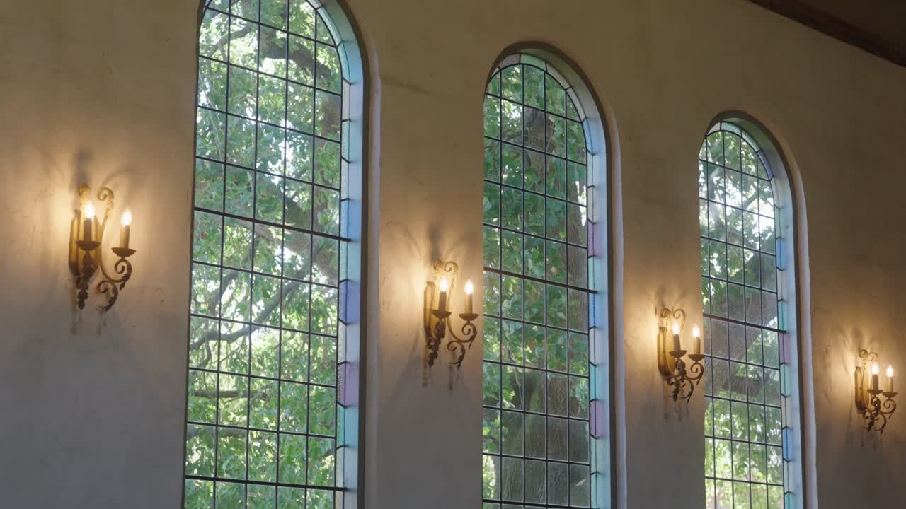 Interior shot of a vintage arched church window with warm wall lamp on the side and a leafy tree visible outside, creating a peaceful and nostalgic atmosphere