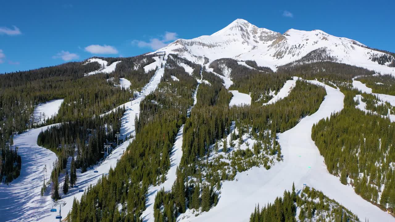 Drone soars over Big Sky ski resort, capturing a busy day of skiing with chair lifts in motion, downhill skiers on snowy trails, and pine-covered mountains bathed in crisp winter light