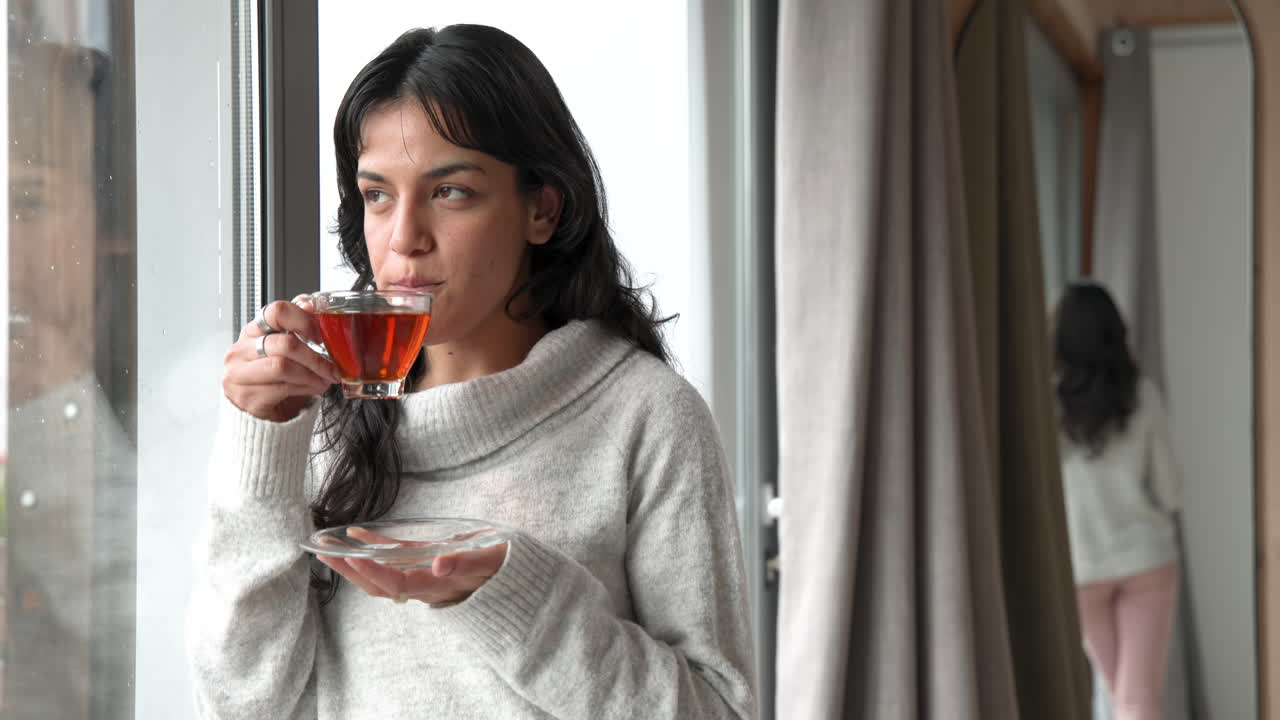 Woman enjoying hot tea by window, wearing cozy sweater, feeling relaxed