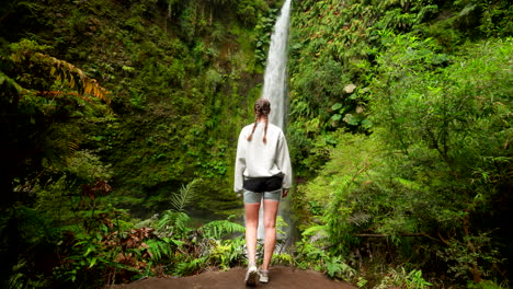 a young woman wearing a sweater and shorts walks through tropical jungle to reach a hidden waterfall in a remote, green forest. concept of eco-tourism, solitude, and outdoor exploration
