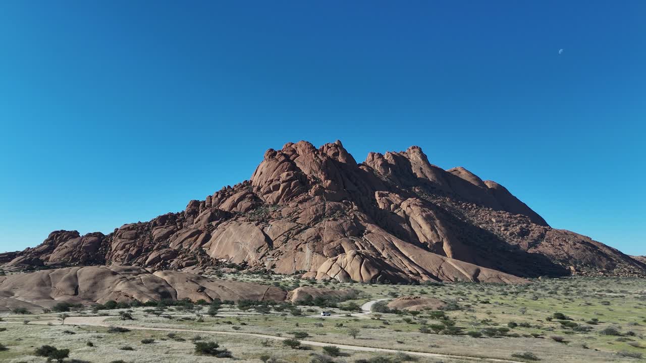 Aerial view of Spitzkoppe mountain in Namibia, showing dramatic granite peaks rising from the desert under clear blue skies, surrounded by arid wilderness