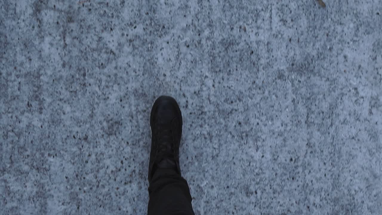 Top down POV or point of view of person with dark clothes walking on a winter road covered in ice, snow and small pebbles that are meant to stop slipping. Movement is smooth, filmed during cloudy day.