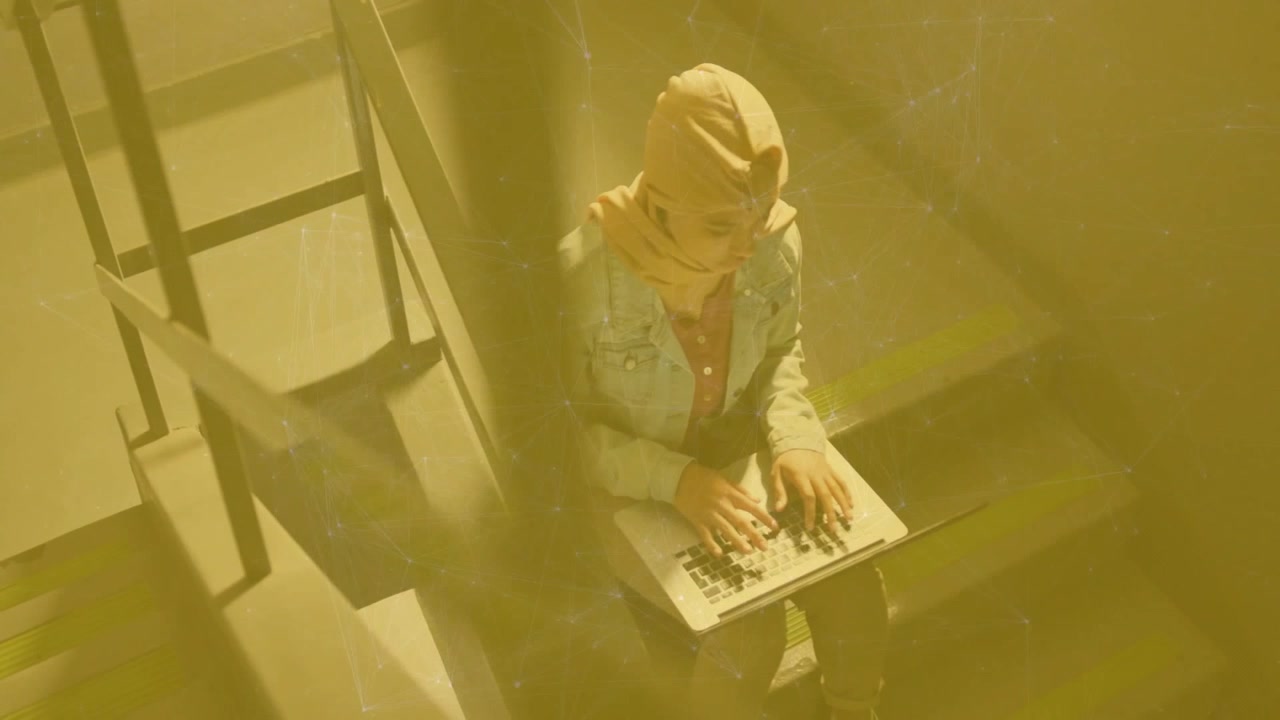 Woman wearing headscarf typing on laptop on stairway through golden tinted barrier for tech work