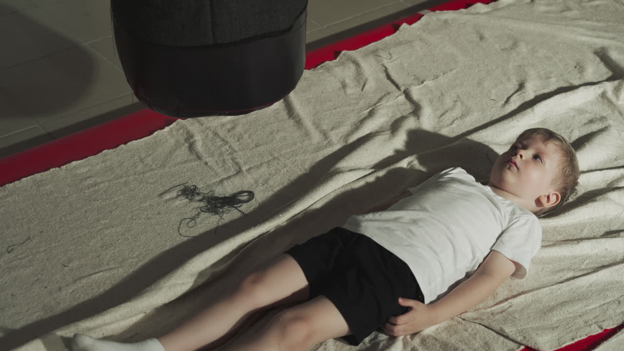 아이는 휴식 시간 동안 권투 체육관에서 카에 앉아 있습니다. 심각한 유아 소년은 전투 스포츠 클럽에서 싱 가방 근처에 누워 있습니다. 아이는 몸을 편안하게합니다.